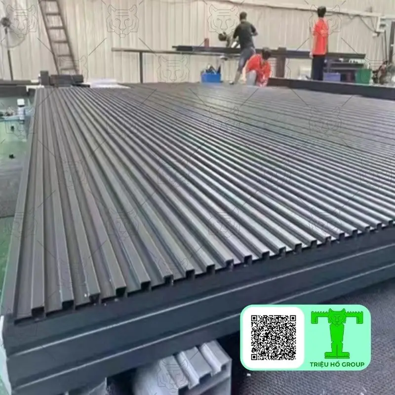Chồng tấm lợp hợp kim nhôm có sóng màu xám đậm trong nhà xưởng, phía sau có công nhân đang làm việc