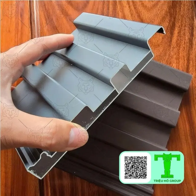 Bàn tay đang cầm một mẫu tấm lợp hợp kim nhôm có cấu trúc sóng, màu xám ở mặt ngoài và nâu ở mặt trong
