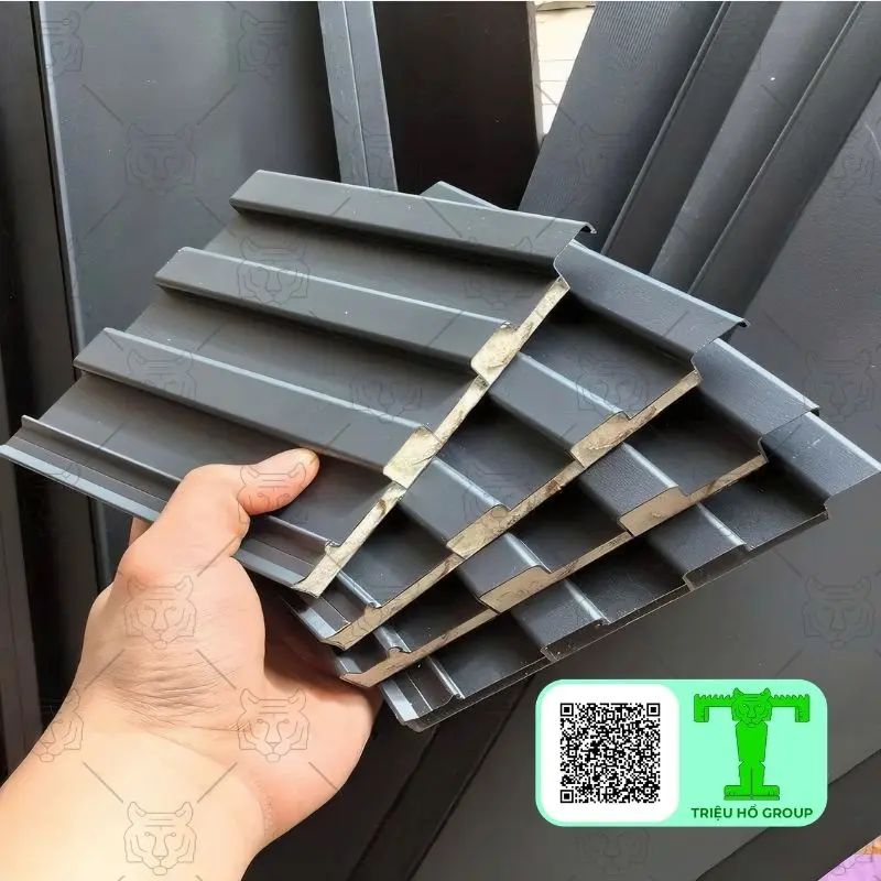 Bàn tay đang cầm nhiều tấm lợp hợp kim nhôm màu xám đậm có sóng, cho thấy cấu trúc vật liệu bên trong