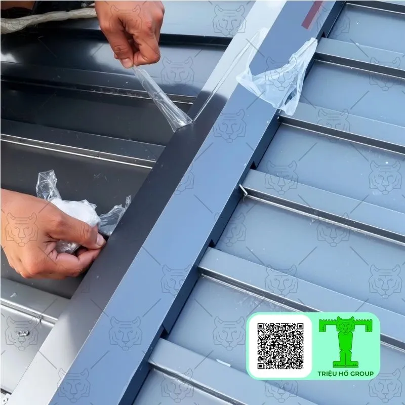 Hai bàn tay đang bóc lớp màng bảo vệ khỏi tấm lợp kim loại màu xám đậm có rãnh, cho thấy bề mặt hoàn thiện